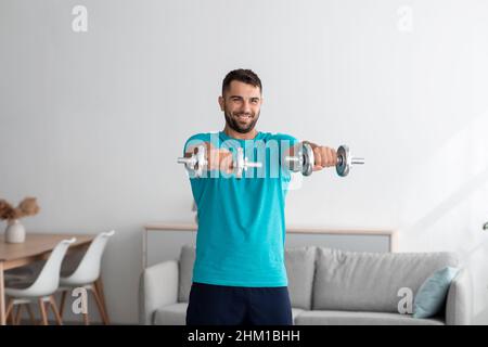 Felice allegro uomo europeo muscoloso millennial facendo esercizi sul braccio, solleva manubri, allenamento per bicipiti Foto Stock