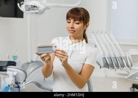 Il dentista dimostra la guida delle tonalità di colore dei denti per sbiancare i denti. Primo piano dei campioni di corrispondenza dei colori dei denti per impianti o impiallacciature nelle mani del medico. Foto di concetto sul tema dentale. Foto Stock