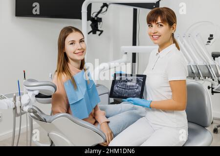 Dentista amichevole con immagine radiografica sulla tavoletta che spiega il trattamento al paziente durante l'esame dei denti. Moderna clinica dentale. Concetto di cura orale professionale, consultazione in studio di stomatologia. Foto Stock