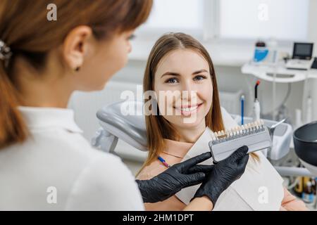 Il dentista dimostra per il paziente le sfumature di colore guida per denti sbiancanti. Campioni di corrispondenza del colore dei denti per impianti o impiallacciature nelle mani del medico. Foto di concetto sul tema dentale. Foto Stock