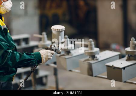 Rivestimento con primer per polveri di parti metalliche. L'uomo di lavoro in un vestito protettivo spruzza la vernice in polvere dalla pistola sulla costruzione del prodotto metallico nello stabilimento di fabbrica. Foto Stock