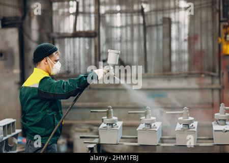 Rivestimento con primer per polveri di parti metalliche. L'uomo di lavoro in un vestito protettivo spruzza la vernice in polvere dalla pistola sulla costruzione del prodotto metallico nello stabilimento di fabbrica. Foto Stock