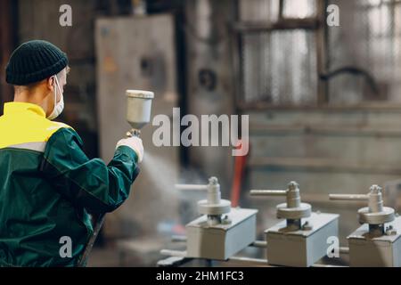 Rivestimento con primer per polveri di parti metalliche. L'uomo di lavoro in un vestito protettivo spruzza la vernice in polvere dalla pistola sulla costruzione del prodotto metallico nello stabilimento di fabbrica. Foto Stock