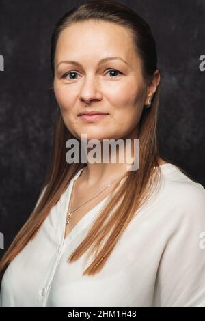 Giovane donna con lunghi capelli biondi e una blusa bianca che si posa di fronte ad uno sfondo grigio scuro. Foto Stock