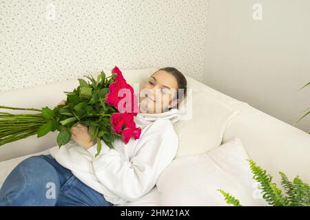 Bella ragazza felice si trova sul letto con un bouquet di rose e li guarda Foto Stock