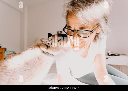 Una vecchia donna con bicchieri sorridenti e baciando il suo gatto sulla sua camera da letto durante una giornata super luminosa Foto Stock