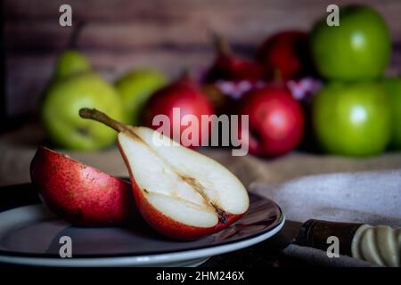 Una pera rossa tagliata a metà su un piatto con più pere e mele fuori fuoco sullo sfondo. Prodotti biologici e naturali. Cibo sano. Foto Stock