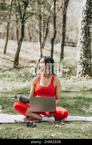 Giovane donna in abiti fitness rilassarsi sul suo computer portatile afferrare una bottiglia d'acqua al parco su un tappetino yoga Foto Stock