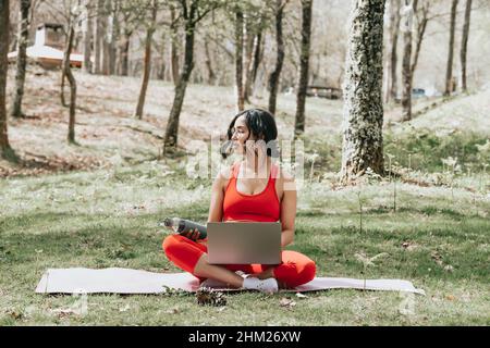 Giovane donna in abiti fitness rilassarsi sul suo computer portatile afferrare una bottiglia d'acqua al parco su un tappetino yoga Foto Stock