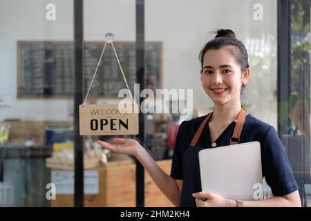 Bella donna giovane barista asiatica in grembiule con tavoletta e in piedi di fronte alla porta del caffè con cartello aperto. SME di avvio del proprietario dell'azienda Foto Stock