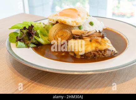 Primo piano Burger giapponese e curry con riso bianco, condito con formaggio, funghi Shimeji, uova fritte. Cibo su un piatto bianco rotondo con insalata fresca. Foto Stock