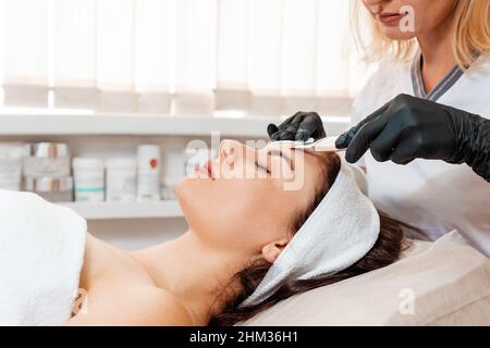 Cosmetologia professionale. Il cosmetologo in guanti di lattice pulisce il viso del cliente utilizzando una macchina a ultrasuoni. Vista laterale. Concetto di procedure di bellezza. Foto Stock