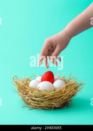 Mano della donna che raggiunge per un uovo rosso di Pasqua su uno sfondo verde-menta. Uova di pollo rosse e bianche in un nido fatto da paillette su un tavolo verde. Foto Stock