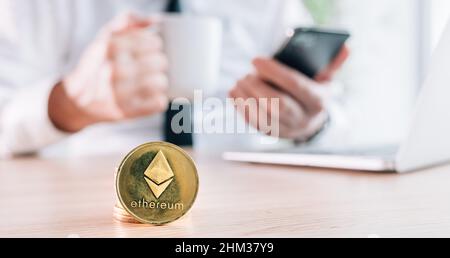 Uomo d'affari che commercia con Ethereum Cryptocurrency piattaforma di commercio sopra il suo smartphone e computer portatile in ufficio, monete impilate davanti, selectiv Foto Stock