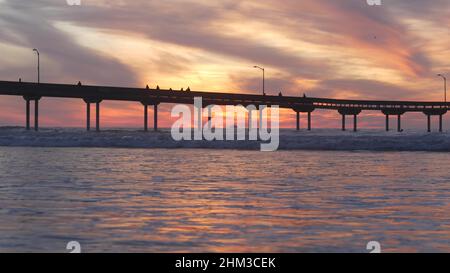 Silhouette di persone che camminano, molo su pali in acqua di mare. Onde oceaniche, cielo drammatico al tramonto. California costa estetica, spiaggia o spiaggia vibrazione al tramonto. Estate stagcape a San Diego vicino Los Angeles Foto Stock