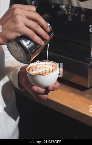 Primo piano mano di uomo barista versare il latte in schiuma in caffè espresso in tazza di caffè rendendo caffe latte art servire al cliente. Piccolo business cafe e res Foto Stock