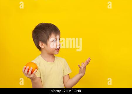 Un bambino allegro in una T-shirt gialla su sfondo giallo tiene un arancione nelle sue mani. Il concetto di cibo sano per i bambini. Spazio di copia Foto Stock