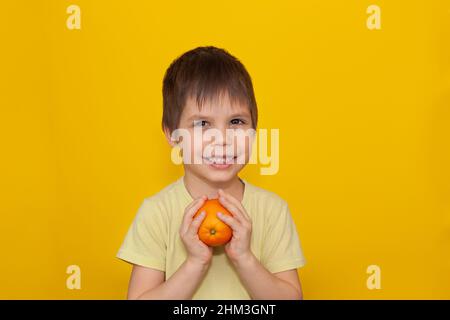 Un bambino allegro in una T-shirt gialla su sfondo giallo tiene un arancione nelle sue mani. Il concetto di cibo sano per i bambini. Spazio di copia Foto Stock