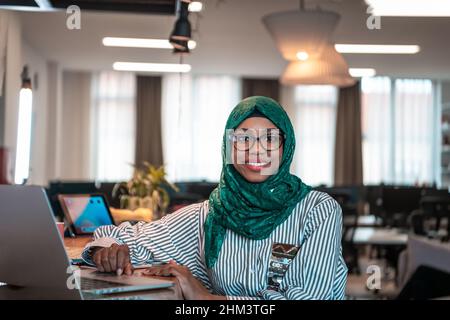 Donna d'affari che indossa un hijab verde utilizzando un computer portatile in zona relax al moderno open plan startup Office. Messa a fuoco selettiva Foto Stock