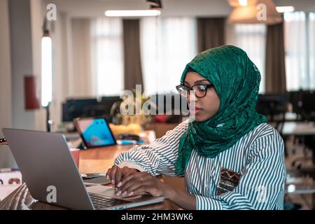 Donna d'affari che indossa un hijab verde utilizzando un computer portatile in zona relax al moderno open plan startup Office. Messa a fuoco selettiva Foto Stock