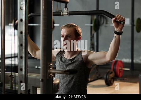 Il giovane atleta concentrato che fa l'esercitazione nella macchina di pull-down in palestra per lavorare i muscoli sui lati del suo petto e della parte superiore della schiena Foto Stock