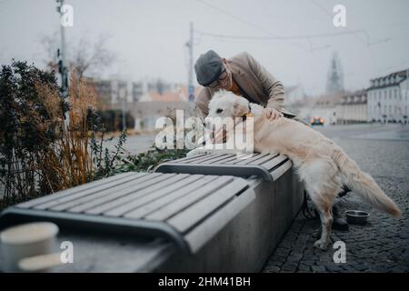 Uomo anziano felice seduto su panca e riposato durante la passeggiata del cane all'aperto in città. Foto Stock