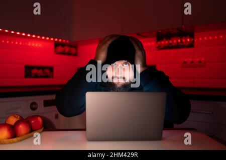 un hacker in una felpa nera con cappuccio sulla testa è seduto su un computer portatile con le mani dietro la testa Foto Stock