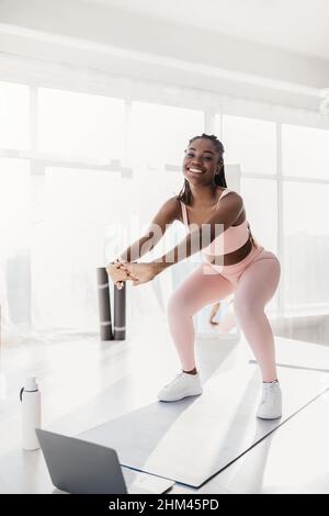 Allenamento online per la forza domestica. Motivato giovane donna nera che fa squats vicino al PC portatile in casa Foto Stock