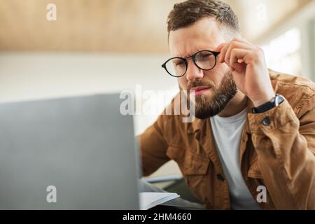 Uomo che guarda il laptop che sbatte gli occhi indossando occhiali da interno Foto Stock