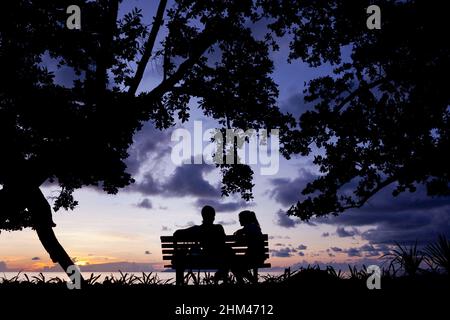 Una coppia che prende il tramonto a Beau Vallon Beach da una panchina parco sull'isola di Mahe, Seychelles. Foto Stock