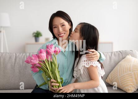 Carina ragazza asiatica baciare la nonna allegra, dando i suoi fiori per il giorno delle Donne, saluto con compleanno a casa Foto Stock