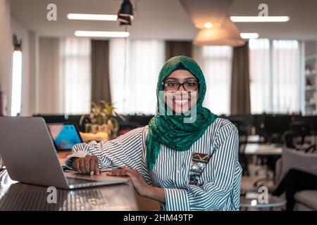 Donna d'affari che indossa un hijab verde utilizzando un computer portatile in zona relax al moderno open plan startup Office. Messa a fuoco selettiva Foto Stock