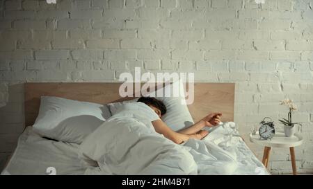 uomo che dorme sotto coperta bianca vicino alla sveglia vintage e orchidea sul comodino Foto Stock