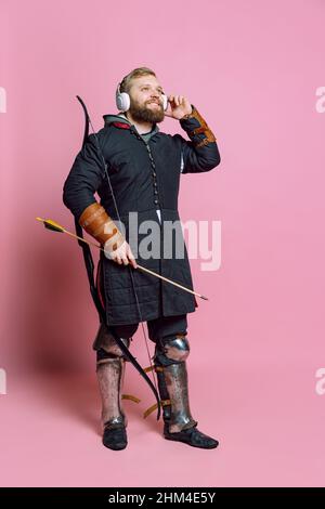 Ritratto di giovane uomo portico in immagine di guerriero medievale, arciere in armatura a catena ascoltando musica in cuffie isolate su sfondo rosa Foto Stock