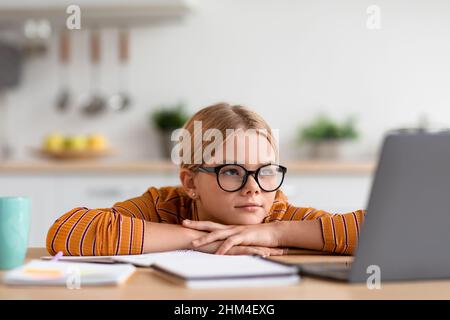 Seria teenage caucasica ragazza bionda in occhiali si siede al tavolo con laptop, guarda video lezione in cucina minimalista Foto Stock