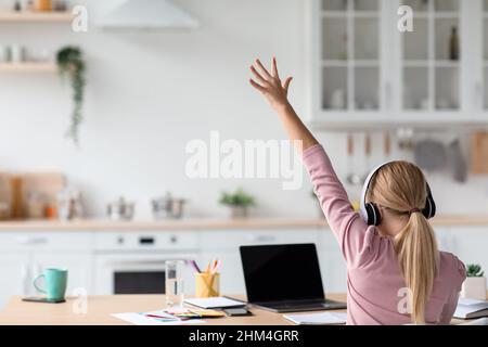 Caucasica teen girl bionda in cuffie studio a casa, alza la sua mano per risposta guarda il pc con schermo vuoto Foto Stock