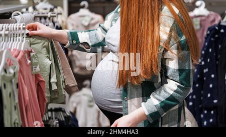 Primo piano di pancia di donna incinta dai capelli rossi nel negozio di cose per bambini, centro commerciale, stagione di sconti e vendite. La madre in attesa sceglie il coagulo Foto Stock