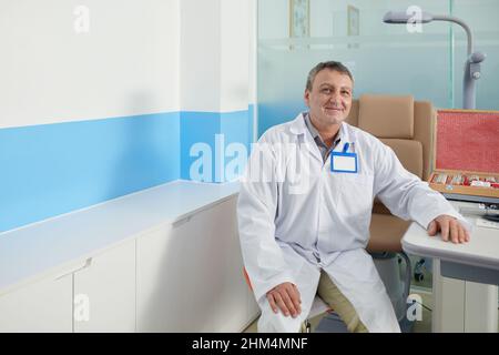 Ritratto di felice optometristo anziano seduto al tavolo nel suo ufficio pronto per esaminare la vista di pazienti con vari strumenti e attrezzature Foto Stock