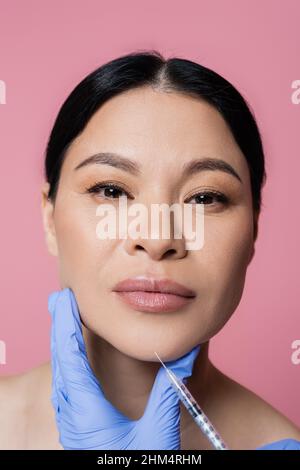 Cosmetologist che tiene la siringa vicino al mento della donna asiatica guardando la macchina fotografica isolata su rosa Foto Stock