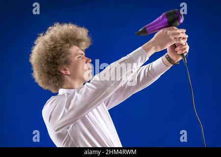 Umorismo. Per asciugare i capelli. Capelli bellissimi. Shaggy ragazzo. Divertente studente. Dirigete l'aria verso di voi. Bell'uomo dai capelli rossi che tiene un asciugacapelli nella sua ha Foto Stock