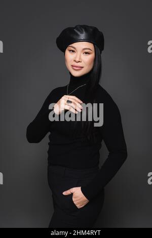 Elegante donna asiatica in beretta in posa isolata su grigio scuro Foto Stock