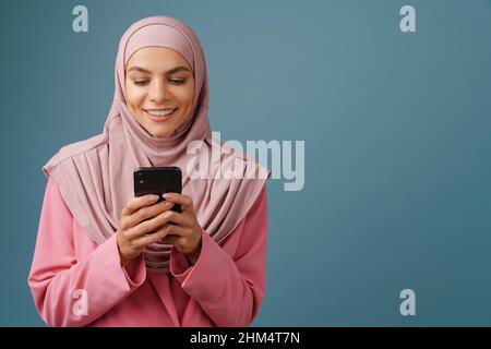Giovane donna musulmana in hijab sorridente mentre usando il telefono cellulare isolato su sfondo blu Foto Stock