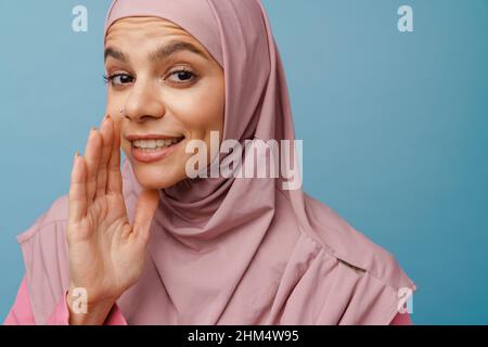 Giovane donna musulmana in hijab gesturing mentre sussurra isolato su sfondo blu Foto Stock