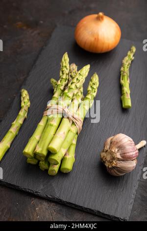 Mazzo di asparagi verdi freschi, aglio, cipolla su tavola di ardesia su fondo di cemento nero. Vista laterale, primo piano. Raccolto, sano, cibo vegano, concetto. Foto Stock