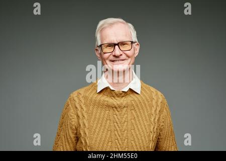 Allegro uomo grigio invecchiato con capelli che indossa maglione marrone lavorato a maglia e occhiali in piedi su sfondo grigio Foto Stock