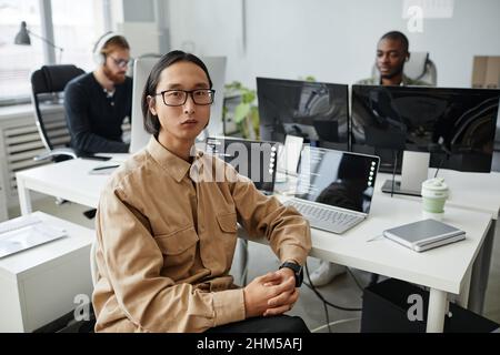 Giovane uomo d'affari asiatico serio in occhiali e abiti casual seduti da scrivania con due computer portatili contro i suoi colleghi Foto Stock