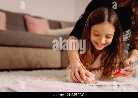 Felice sorridente bambina passare il tempo con sua madre che si trova sul tappeto a casa e disegnando sulla carta immagine. Foto Stock