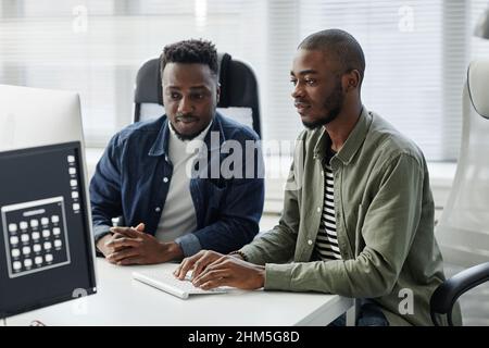 Due giovani programmatori contemporanei di diversità in abiti casual che immettono dati nel computer mentre si siedono alla scrivania durante la riunione Foto Stock