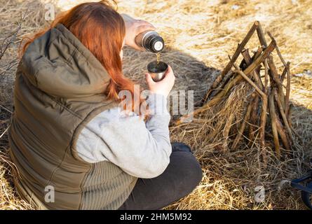 Foto dall'alto di redhair viaggiatore donna vestito calorosamente con falò e versando il tè caldo da thermos bottiglia nella tazza. Foto Stock