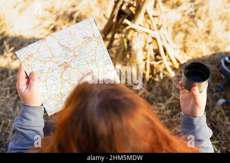 Vista dall'alto foto di una singola donna viaggiatore con redhairs seduta di fronte al falò mantenendo nelle sue mani una tazza di metallo di tè caldo e uno sguardo Foto Stock